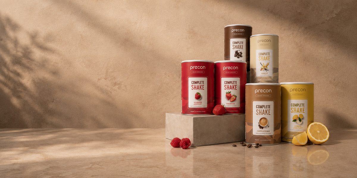 Precon Complete Shakes in verschiedenen Sorten, gestapelt auf einem Podest mit Himbeeren und Zitronen