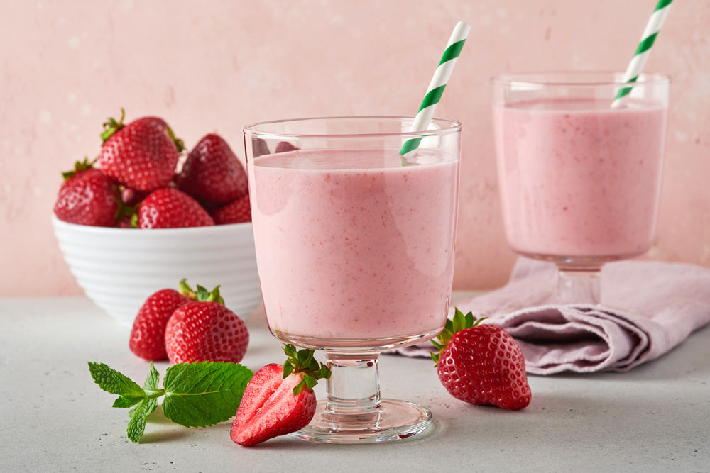 Zwei Gläser Erdbeer-Smoothie mit grün gestreiften Strohhalmen auf einer hellen Fläche, garniert mit frischen Erdbeeren und Minze, im Hintergrund eine Schale mit Erdbeeren.