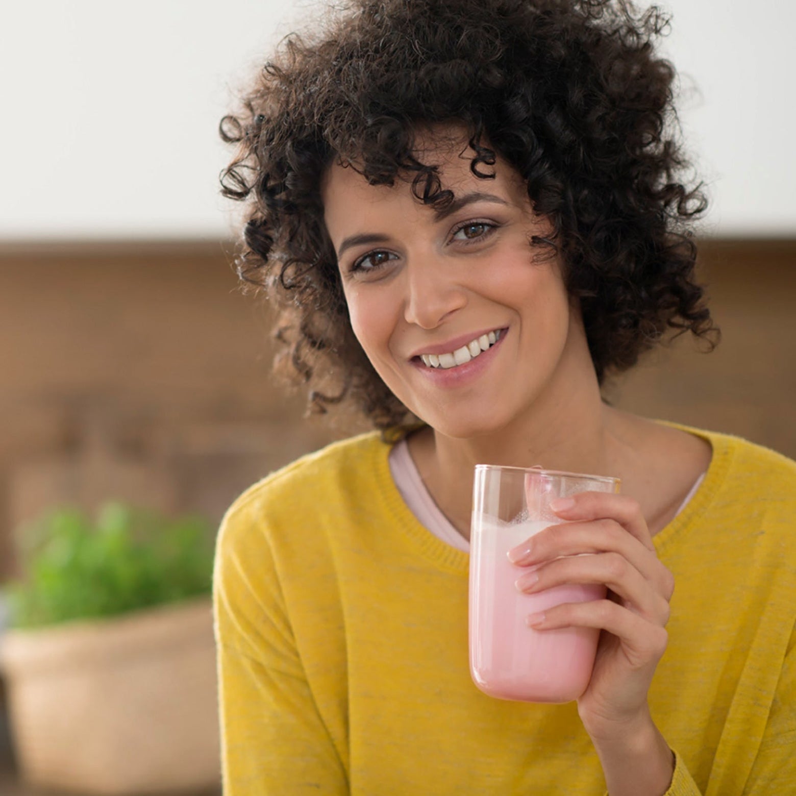 Eine Frau mit lockigem Haar im gelben Pullover lächelt, in der Hand ein Glas rosa Precon Diät Smoothie aus dem Einsteiger-Startpaket CH. Ein sanft verschwommener Hintergrund zeigt eine gemütliche Küche.