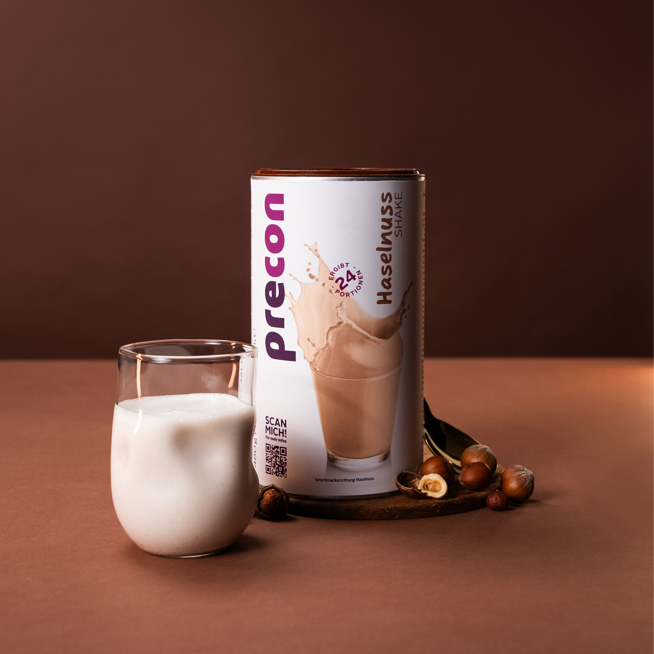 Ein cremiges Glas Precon Haselnuss-Shake steht neben seinem Behälter auf einer braunen, mit Haselnüssen bestreuten Fläche vor einem Hintergrund mit warmem Braun.
