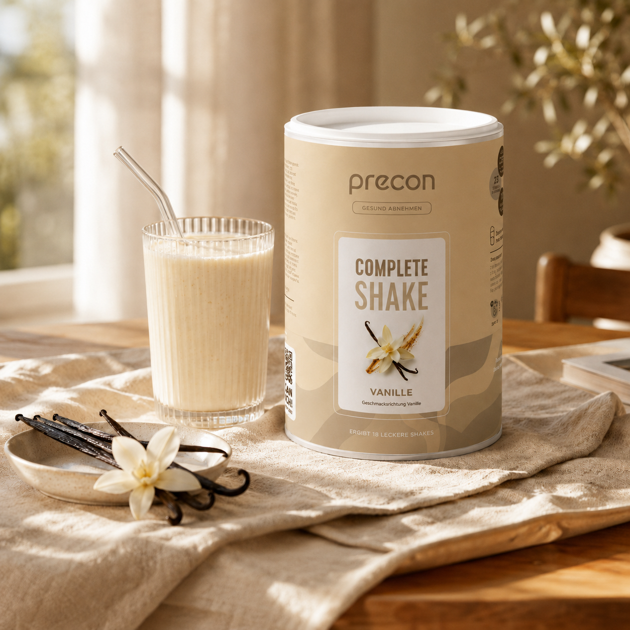 Ein Glas Vanille-Shake mit einem Strohhalm steht neben einer Packung „Complete Shake Vanille“-Pulver der Anona GmbH, zusammen mit Vanilleschoten und einer Vanilleblüte auf einer hellen Tischdecke in einem sonnendurchfluteten Raum.