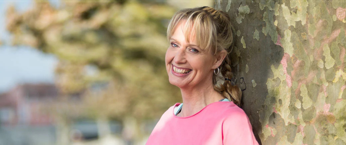 Lachende Frau in pinkem Shirt lehnt entspannt an einem Baumstamm – gelassen mit Ausrutschern umgehen
