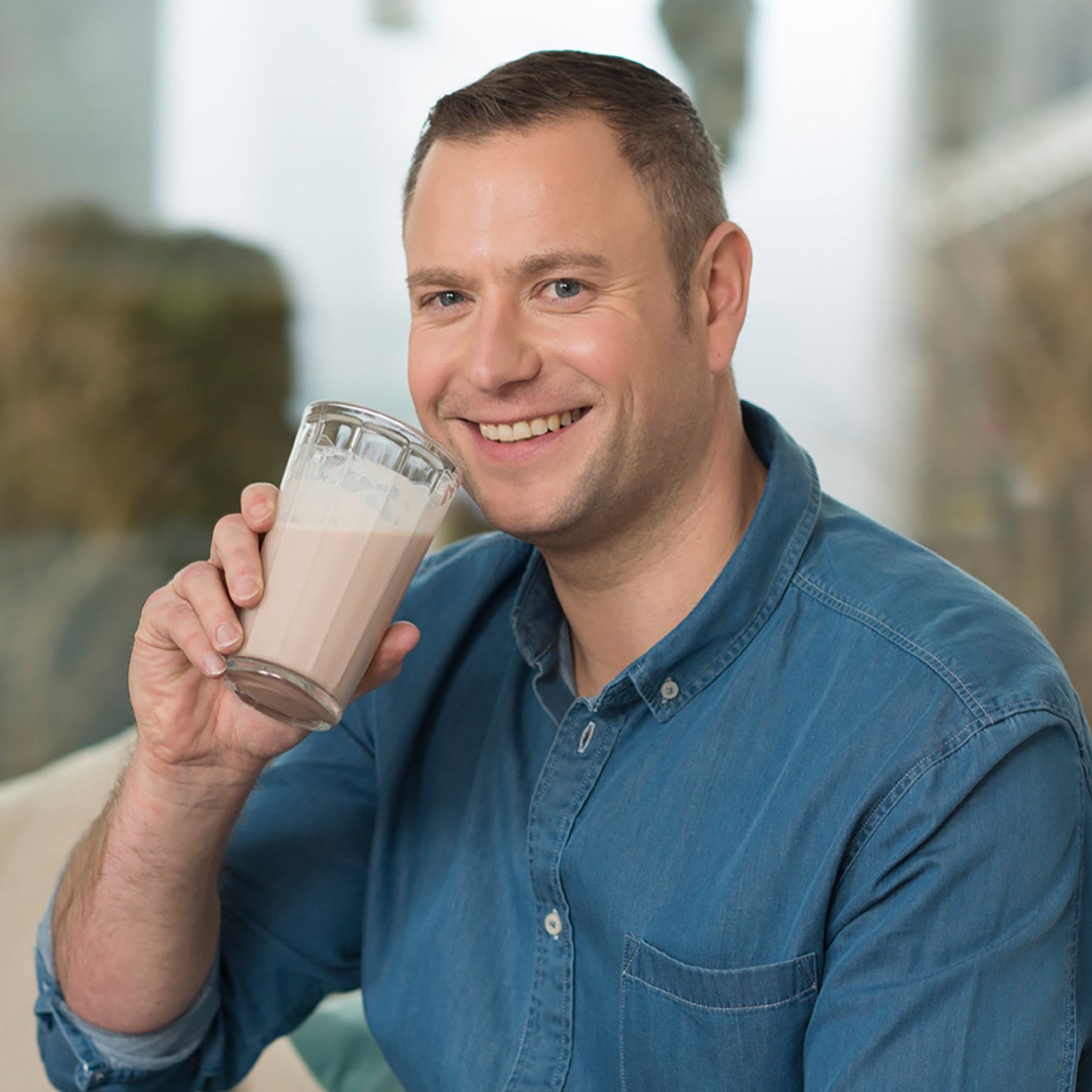 Ein lächelnder Mann in einem blauen Jeanshemd hält ein Glas Precon Start Shake Schoko-Schokoladenmilch in der Hand, während er in einem Innenraum in einer hellen, sanft konzentrierten Umgebung sitzt.