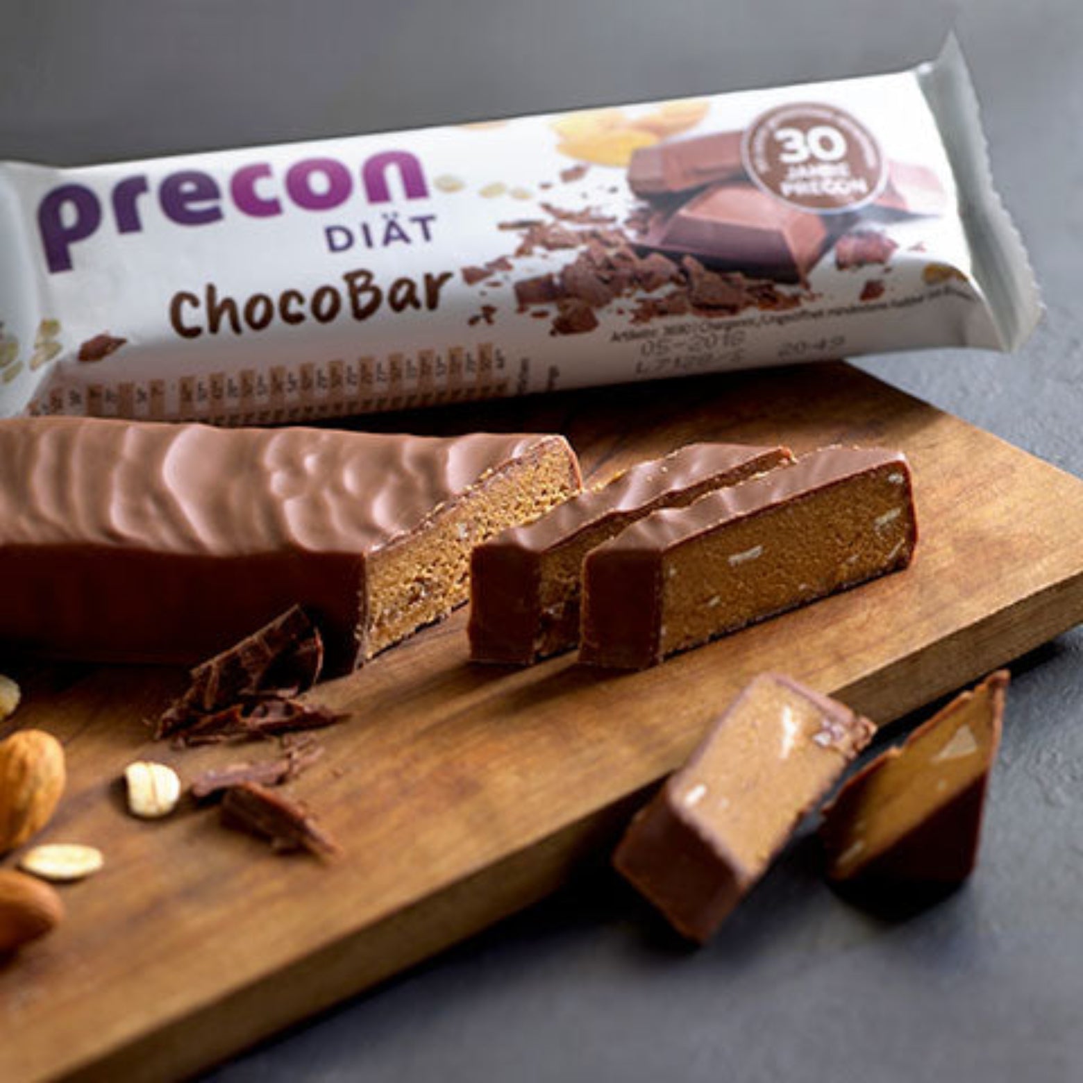 Ein aufgeschnittener Precon ChocoBar Riegel wird auf einem Holzbrett mit Mandeln und Hafer neben der Verpackung mit der Aufschrift "Precon ChocoBar - 30% Protein" gezeigt, um diesen köstlichen Mahlzeitenersatz hervorzuheben.